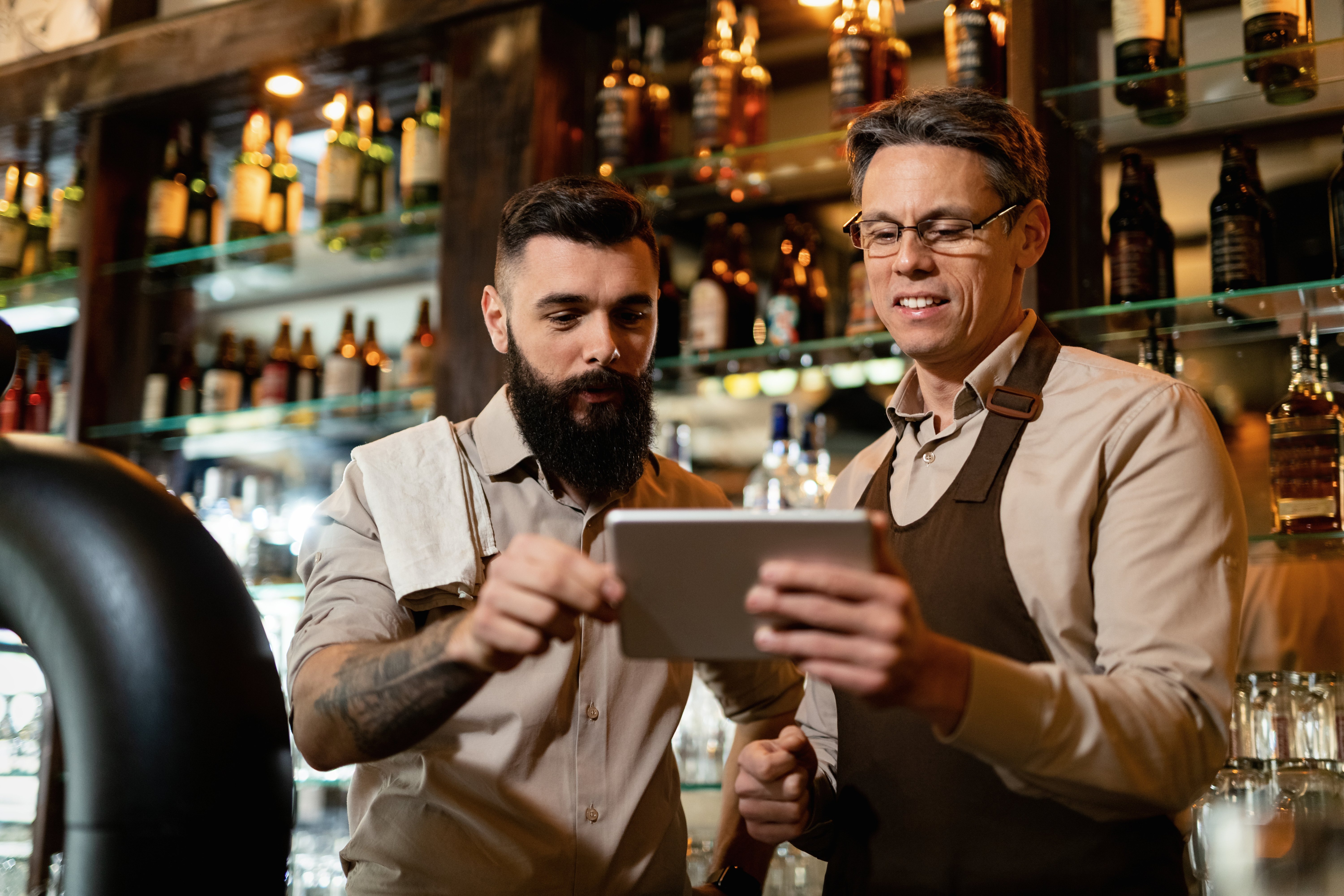 mid-adult-barista-his-young-coworker-using-touchpad-while-working-pub